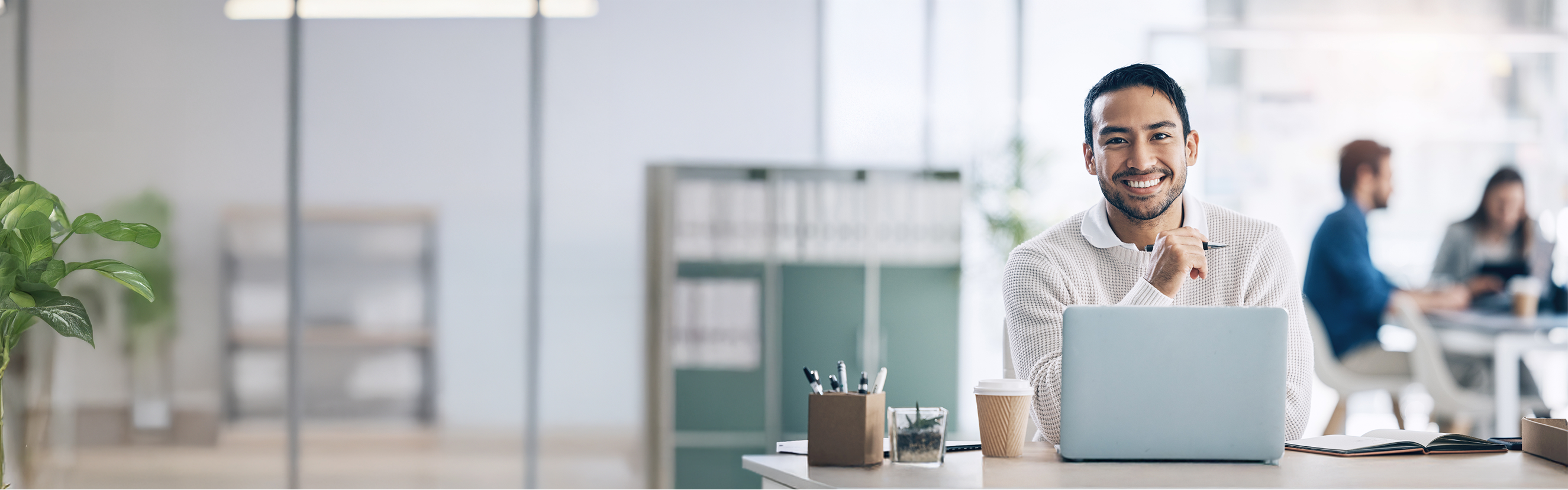 Ein Mann sitzt an einem Schreibtisch in einem modernen Büro und arbeitet an einem Laptop. Im Vordergrund befinden sich Büromaterialien und ein Kaffeebecher. Im Hintergrund sind weitere Personen zu sehen, die ebenfalls arbeiten.