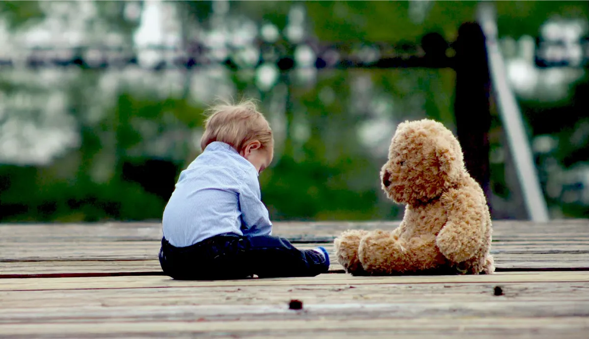 Ein Bild von einem kleinen Kind, das neben seinem Teddybären sitzt.