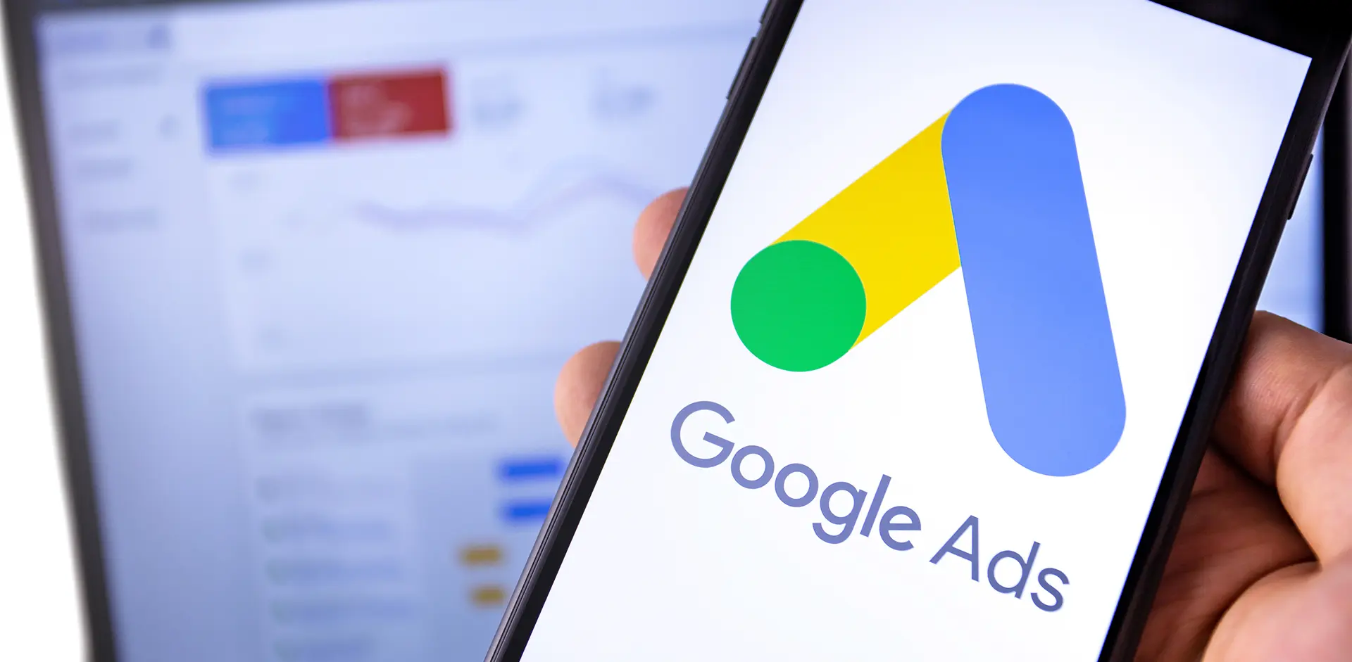 Smartphone mit geöffnetem Google Ads Logo, im Hintergrund unscharfer Bildschirm mit Analytics-Dashboard – Symbol für Online-Werbung und Performance-Marketing.