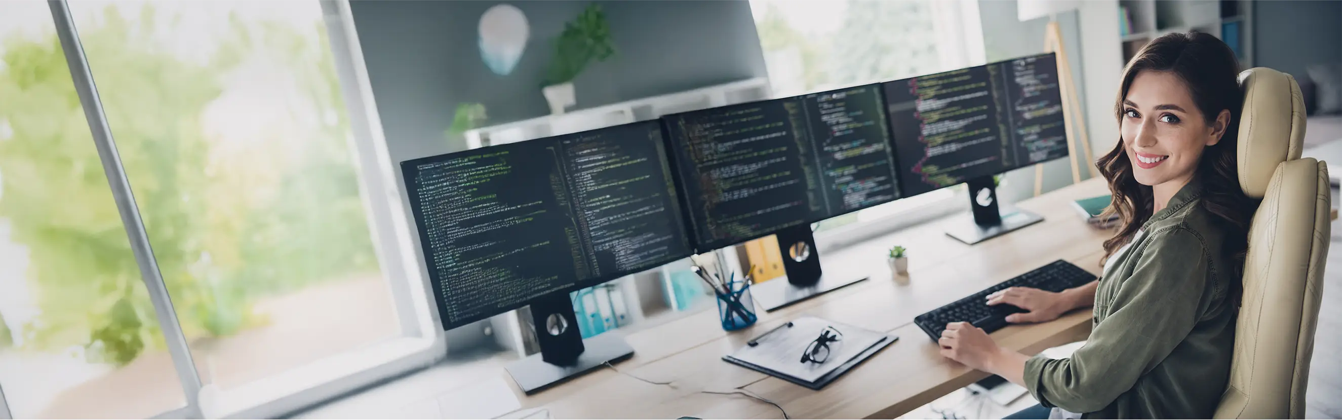 Moderne Arbeitsumgebung mit drei Monitoren, auf denen Code angezeigt wird, und einer lächelnden Entwicklerin am Schreibtisch.