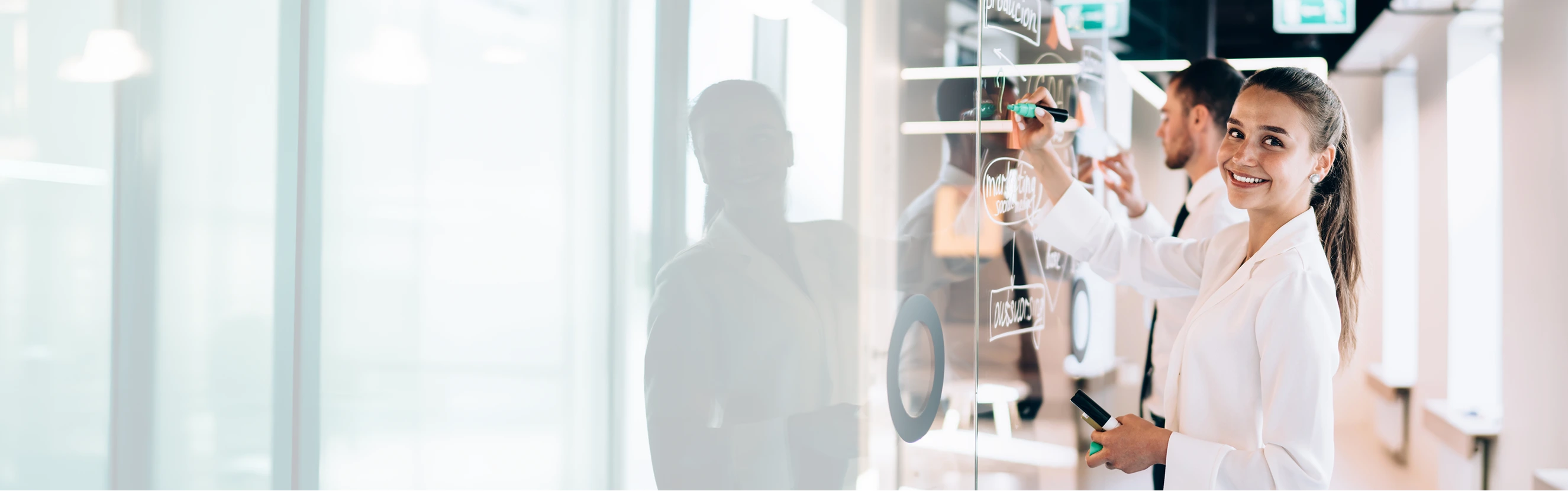 Eine Frau steht vor einer Tafel aus Glas und schreibt mit einem Marker darauf. Sie lächelt in die Kamera. Im Hintergrund arbeitet noch ein weiterer Mann auf der Tafel.