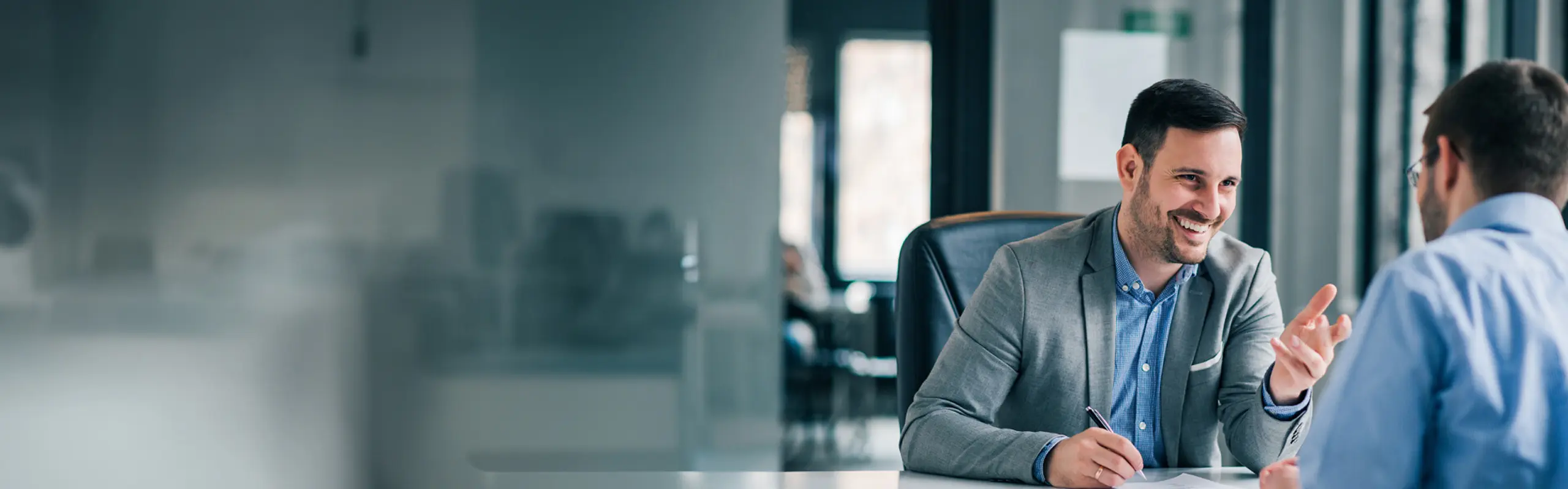 Zwei Männer in einem modernen Büro im Gespräch – einer lächelnd mit Stift in der Hand, der andere mit dem Rücken zur Kamera.